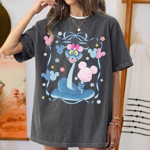 May include: Dark gray t-shirt featuring a cartoon snake with a pink bow, surrounded by pink and blue Mickey Mouse-shaped balloons and flowers. The snake is blue with a pink bow and holding a pink cotton candy.