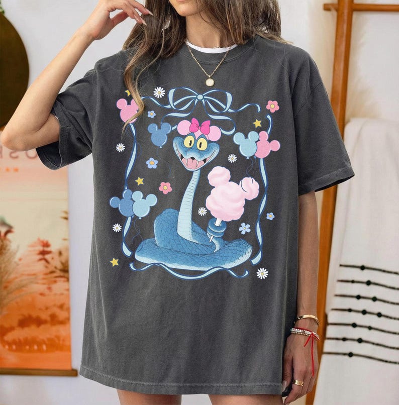 May include: Dark gray t-shirt featuring a cartoon snake with a pink bow, surrounded by pink and blue Mickey Mouse-shaped balloons and flowers. The snake is blue with a pink bow and holding a pink cotton candy.