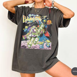 Puede incluir: Una camiseta gris carb&oacute;n de manga corta con un estampado gr&aacute;fico de personajes espaciales. El dise&ntilde;o incluye el texto "ASTRO BLASTERS" en amarillo y azul, con una figura de guardi&aacute;n espacial, alien&iacute;genas verdes y un villano oscuro de fondo.