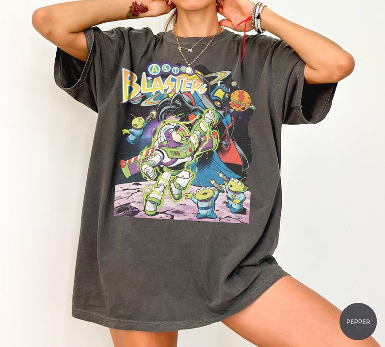 Puede incluir: Una camiseta gris carb&oacute;n de manga corta con un estampado gr&aacute;fico de personajes espaciales. El dise&ntilde;o incluye el texto "ASTRO BLASTERS" en amarillo y azul, con una figura de guardi&aacute;n espacial, alien&iacute;genas verdes y un villano oscuro de fondo.