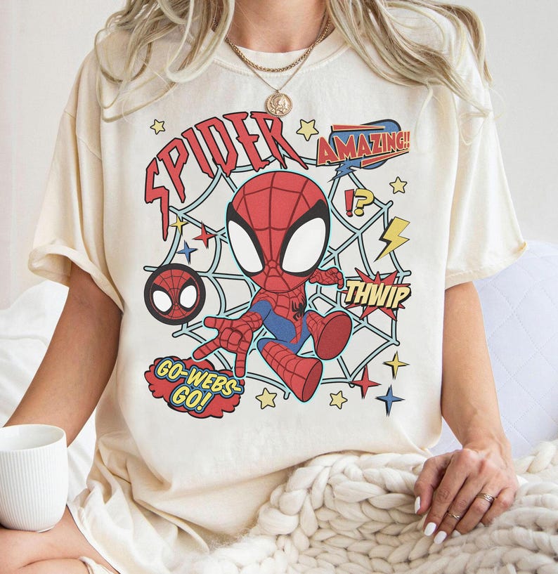 May include: A cream-colored t-shirt featuring a graphic of a cartoon Spider-Man character in a red and blue suit. The design includes a spider web background, yellow lightning bolts, stars, and text that reads SPIDER, AMAZING, THWIP, and GO-WEBS-GO.