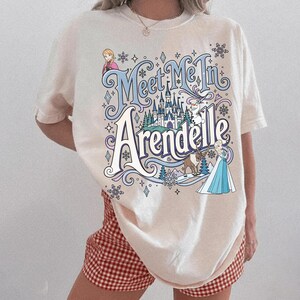 K&ouml;nnte beinhalten: Ein cremefarbenes T-Shirt mit kurzen &Auml;rmeln und einem Grafikdruck mit dem Text "Meet Me In Arendelle" in blauer und wei&szlig;er Schrift. Das Design zeigt Illustrationen von Anna, Elsa, Olaf und Sven, ein Schloss sowie verschiedene Schneeflocken.