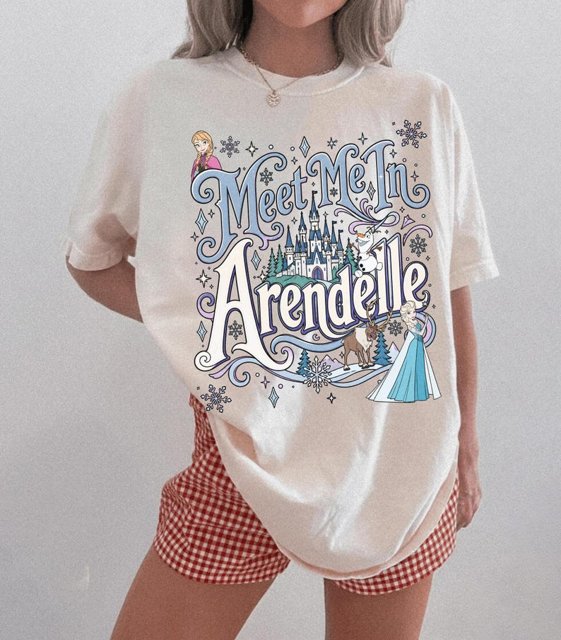 K&ouml;nnte beinhalten: Ein cremefarbenes T-Shirt mit kurzen &Auml;rmeln und einem Grafikdruck mit dem Text "Meet Me In Arendelle" in blauer und wei&szlig;er Schrift. Das Design zeigt Illustrationen von Anna, Elsa, Olaf und Sven, ein Schloss sowie verschiedene Schneeflocken.