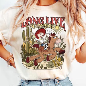 Puede incluir: Una camiseta color crema con un estampado gr&aacute;fico de una vaquera montando un caballo marr&oacute;n con un lazo. El texto dice LONG LIVE cowgirls. El fondo incluye un paisaje des&eacute;rtico con cactus, un sol y plantas en tonos verdes y amarillos.