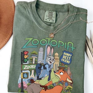 May include: An olive green short-sleeve t-shirt featuring a graphic print of the characters Judy Hopps and Nick Wilde from Zootopia. The text reads Zootopia and Bunny Best Friend. The shirt has a crew neckline and a Comfort Colors brand label.