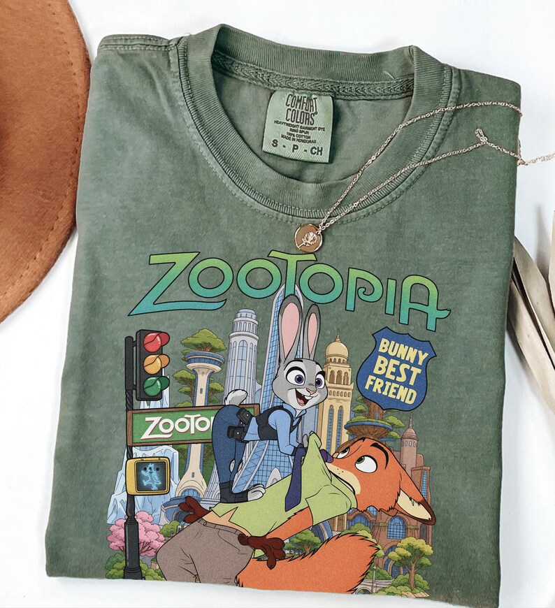 May include: An olive green short-sleeve t-shirt featuring a graphic print of the characters Judy Hopps and Nick Wilde from Zootopia. The text reads Zootopia and Bunny Best Friend. The shirt has a crew neckline and a Comfort Colors brand label.