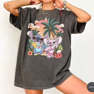 May include: A charcoal gray short-sleeve t-shirt featuring a graphic print of two cartoon characters on a beach. One blue character wears sunglasses and holds a pineapple, while a pink character stands nearby. Palm trees, hibiscus flowers, and a sunset sky complete the scene.