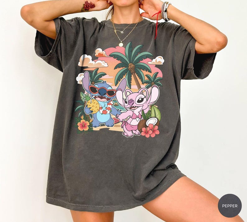 May include: A charcoal gray short-sleeve t-shirt featuring a graphic print of two cartoon characters on a beach. One blue character wears sunglasses and holds a pineapple, while a pink character stands nearby. Palm trees, hibiscus flowers, and a sunset sky complete the scene.