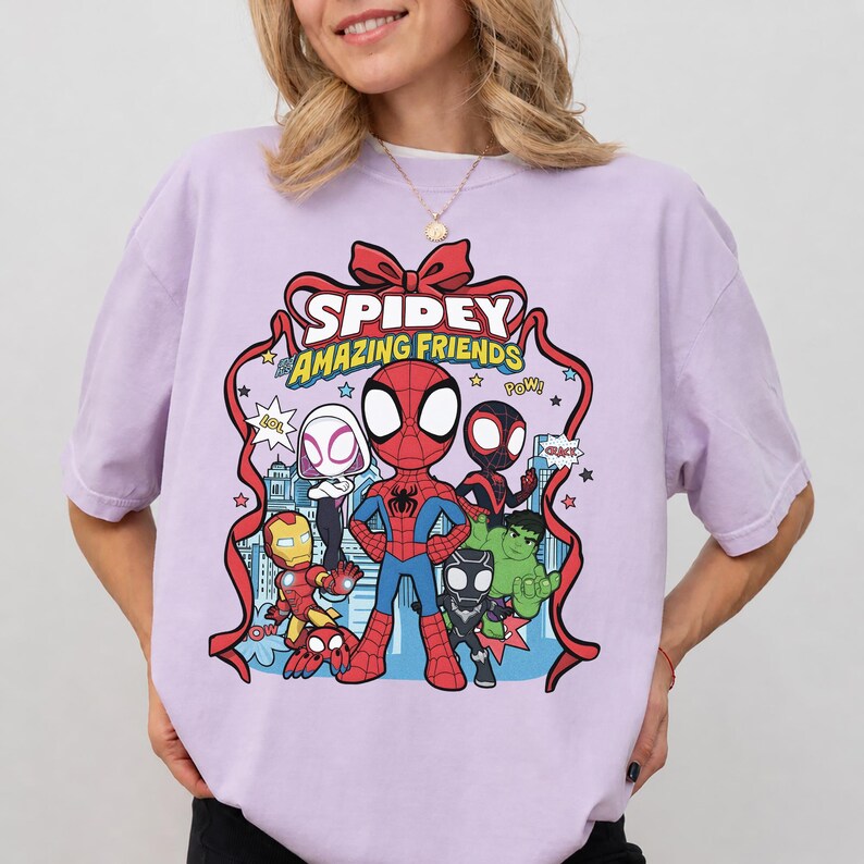 May include: A light purple t-shirt featuring a graphic print of Spidey and His Amazing Friends characters including Spider-Man, Ghost-Spider, Iron Man, Hulk, and Black Panther, framed by a red ribbon with the text Spidey and Amazing Friends.