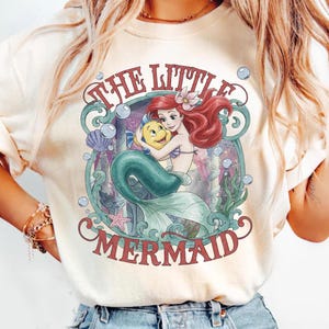 May include: A cream-colored t-shirt featuring a graphic print of Ariel and Flounder from The Little Mermaid. The design includes red text, underwater elements like bubbles, seaweed, a seashell, and a starfish, all framed within a decorative border.