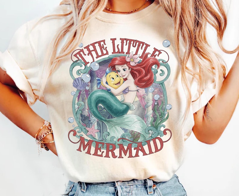 May include: A cream-colored t-shirt featuring a graphic print of Ariel and Flounder from The Little Mermaid. The design includes red text, underwater elements like bubbles, seaweed, a seashell, and a starfish, all framed within a decorative border.