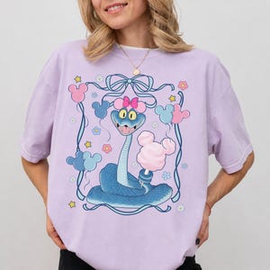 May include: A light purple t-shirt featuring a cartoon snake holding cotton candy. The snake has a pink bow and is surrounded by Mickey Mouse-shaped balloons and flowers. The design is framed by a blue ribbon.