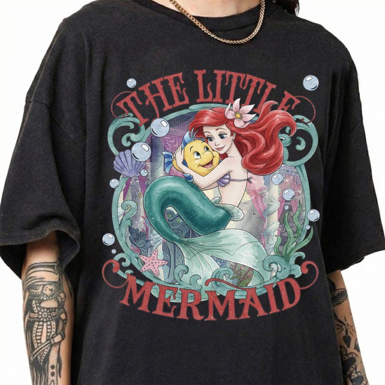 May include: A black short-sleeve t-shirt featuring a graphic print of Ariel and Flounder from The Little Mermaid. The design includes the text THE LITTLE MERMAID, surrounded by underwater elements like bubbles, a starfish, and a seashell in muted tones.