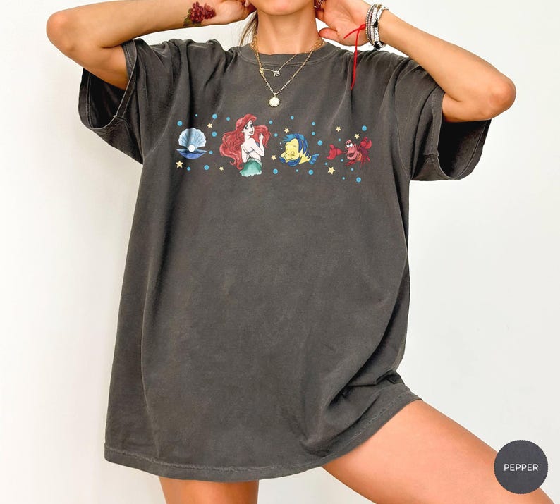 K&ouml;nnte beinhalten: Dunkelgraues T-Shirt mit einem farbenfrohen Aufdruck einer Perle, einer Meerjungfrau, einem Fisch und einer Krabbe. Das Design umfasst blaue Sterne und ist auf einem kurz&auml;rmeligen, &uuml;bergro&szlig;en T-Shirt aufgedruckt. Das Shirt hat einen Vintage-Look.