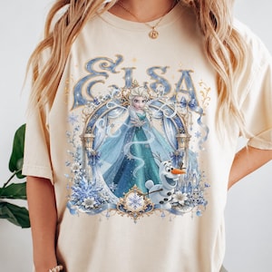 May include: A cream-colored short-sleeve t-shirt featuring a graphic print of Elsa and Olaf from Frozen. The design includes the text Elsa in blue, an ornate archway, snowflakes, and blue ribbons, all centered on the front of the garment.