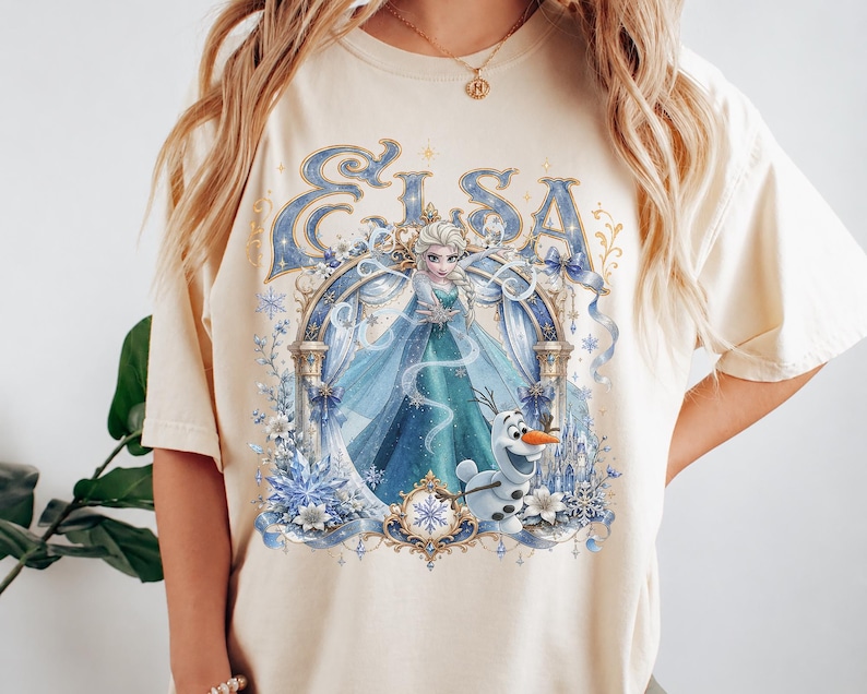May include: A cream-colored short-sleeve t-shirt featuring a graphic print of Elsa and Olaf from Frozen. The design includes the text Elsa in blue, an ornate archway, snowflakes, and blue ribbons, all centered on the front of the garment.