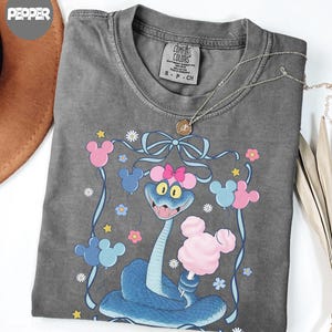 May include: A gray Comfort Colors t-shirt featuring a cartoon snake with a pink bow, holding cotton candy. The design includes Mickey Mouse-shaped balloons in pink and blue, with flowers and stars. A gold necklace is draped across the shirt.
