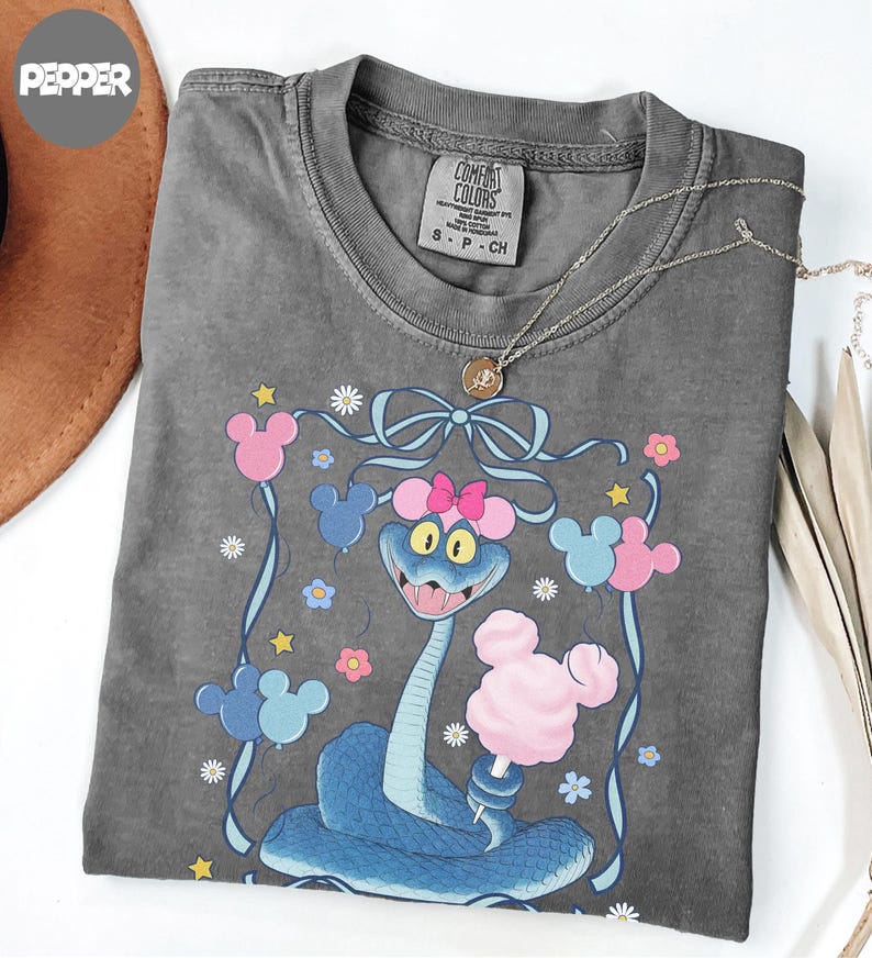 May include: A gray Comfort Colors t-shirt featuring a cartoon snake with a pink bow, holding cotton candy. The design includes Mickey Mouse-shaped balloons in pink and blue, with flowers and stars. A gold necklace is draped across the shirt.