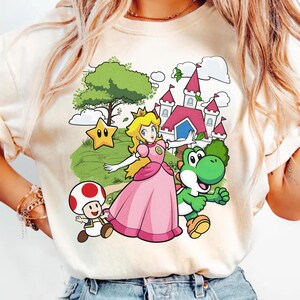 May include: Cream-colored t-shirt featuring a colorful graphic of Princess Peach, Yoshi, and a mushroom character from the Super Mario video game series. The design includes a castle, star, and tree on a green background.