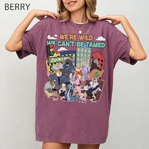 May include: Berry-colored t-shirt featuring a colorful graphic of characters from the animated movie Zootopia. The design includes the text "WE'RE WILD WE CAN'T BE TAMED" and the Zootopia logo. The shirt has short sleeves and a crew neck.