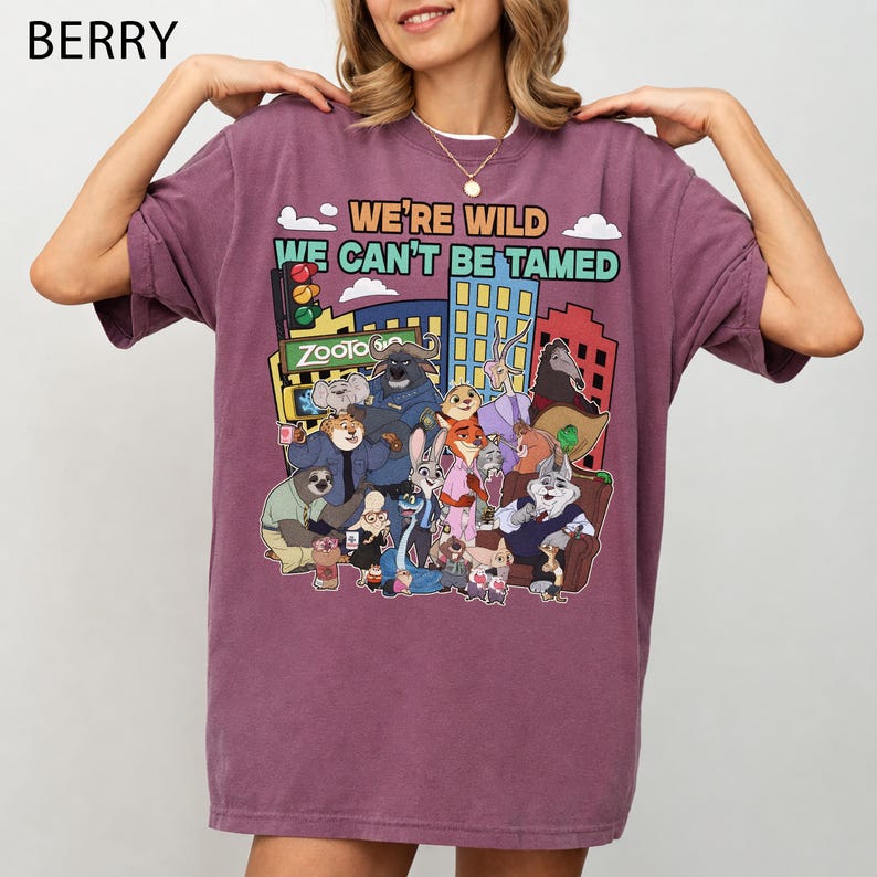 May include: Berry-colored t-shirt featuring a colorful graphic of characters from the animated movie Zootopia. The design includes the text "WE'RE WILD WE CAN'T BE TAMED" and the Zootopia logo. The shirt has short sleeves and a crew neck.