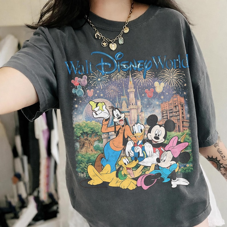 Pu&ograve; includere: T-shirt grigio scuro con una grafica colorata dei personaggi Disney, tra cui Topolino, Minnie, Pippo, Paperino e Pluto, di fronte al castello di Cenerentola. Il testo "Walt Disney World" &egrave; visualizzato in blu.