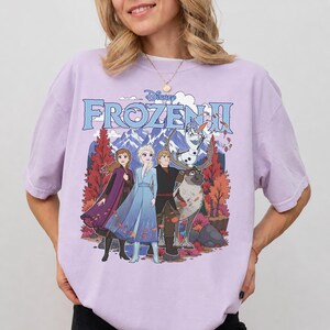 May include: A light purple short-sleeve t-shirt featuring a graphic print of the Disney Frozen II logo above characters Anna, Elsa, Kristoff, Sven, and Olaf standing in front of a mountain landscape with orange and red autumn trees.