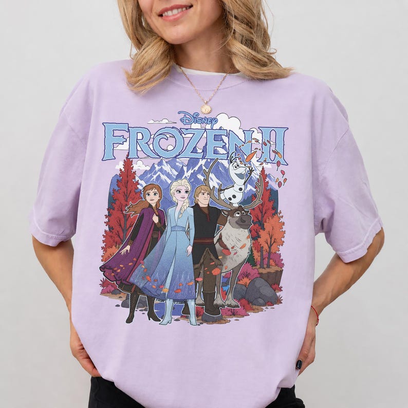 May include: A light purple short-sleeve t-shirt featuring a graphic print of the Disney Frozen II logo above characters Anna, Elsa, Kristoff, Sven, and Olaf standing in front of a mountain landscape with orange and red autumn trees.