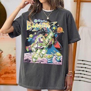Puede incluir: Una camiseta de manga corta gris carb&oacute;n con un estampado gr&aacute;fico de Buzz Lightyear y alien&iacute;genas verdes de Toy Story. El texto Astro Blasters est&aacute; impreso en amarillo y azul sobre los personajes, con un fondo de espacio oscuro.