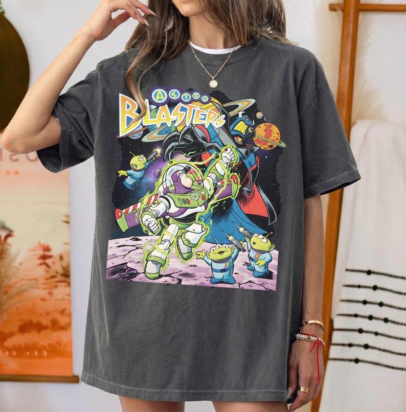 Puede incluir: Una camiseta de manga corta gris carb&oacute;n con un estampado gr&aacute;fico de Buzz Lightyear y alien&iacute;genas verdes de Toy Story. El texto Astro Blasters est&aacute; impreso en amarillo y azul sobre los personajes, con un fondo de espacio oscuro.