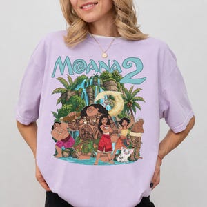 May include: A light purple short-sleeve t-shirt featuring a colorful graphic print of characters from the movie Moana 2. The design includes the text Moana 2 above a tropical island scene with a waterfall, palm trees, and several animated figures.