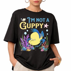 Puede incluir: Camiseta negra con un dise&ntilde;o de dibujos animados de Flounder de La Sirenita y el texto "I'm Not a Guppy". El dise&ntilde;o incluye burbujas, coral y una estrella de mar. Una camiseta c&oacute;moda e informal.