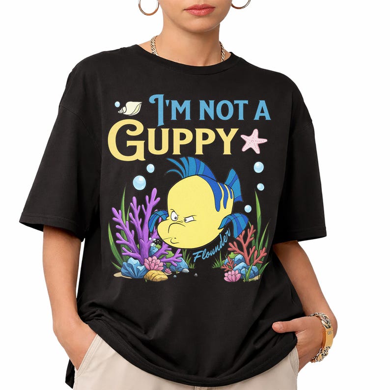 Puede incluir: Camiseta negra con un dise&ntilde;o de dibujos animados de Flounder de La Sirenita y el texto "I'm Not a Guppy". El dise&ntilde;o incluye burbujas, coral y una estrella de mar. Una camiseta c&oacute;moda e informal.