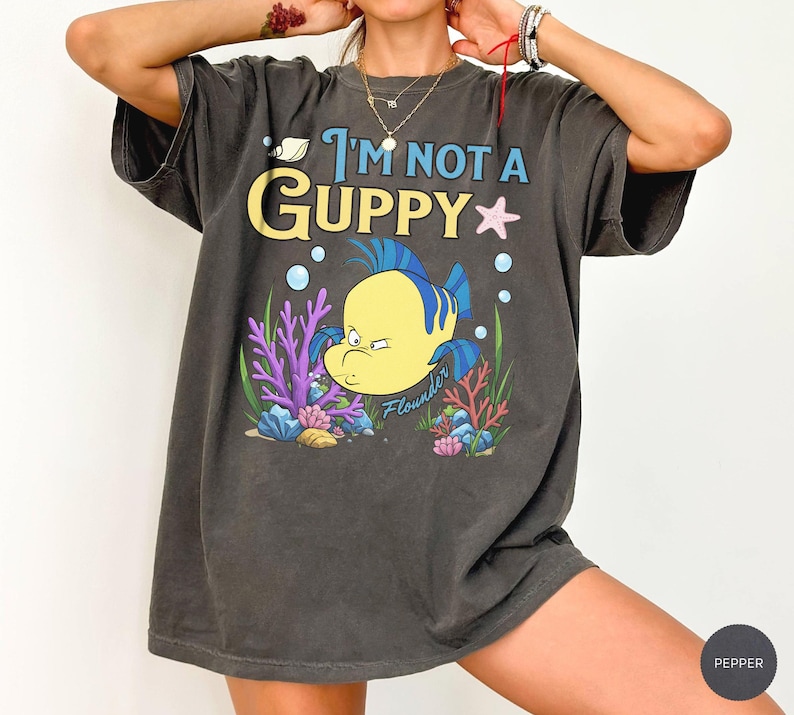 Puede incluir: Camiseta gris oscuro con la frase "I'm Not a Guppy" y una ilustraci&oacute;n de dibujos animados de Flounder de La Sirenita. El dise&ntilde;o incluye un pez amarillo con aletas azules, coral y burbujas.