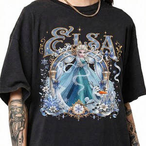 May include: A black short-sleeve t-shirt featuring a graphic print of Elsa and Olaf from Frozen. The design includes the text ELSA in a blue font, surrounded by an ornate blue and gold frame with snowflakes, flowers, and ribbons.