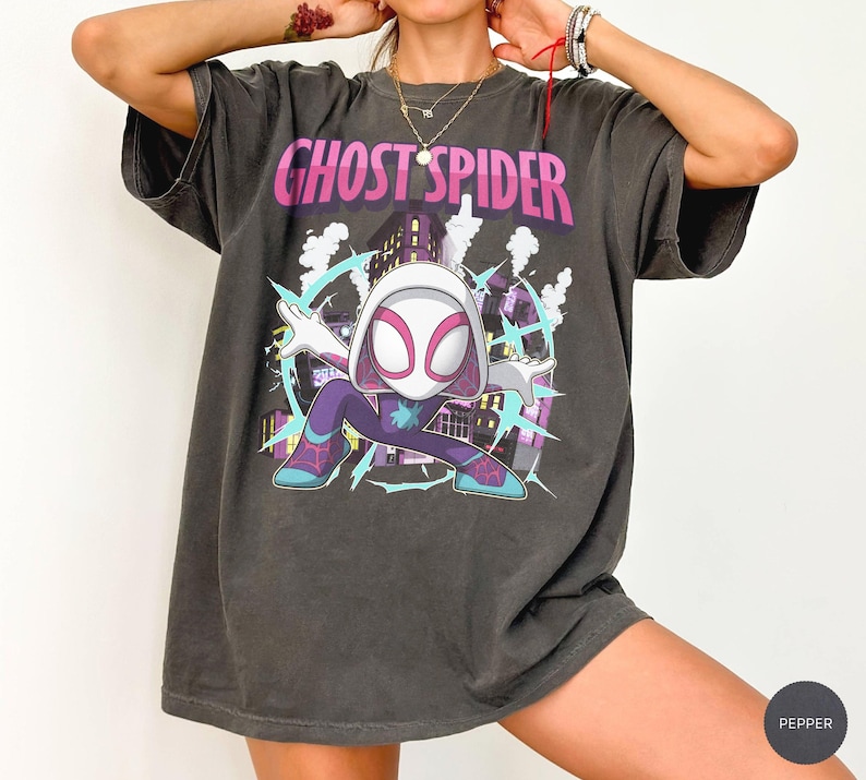 May include: Dark gray t-shirt featuring a cartoon illustration of Ghost Spider. The graphic includes the text "GHOST SPIDER" in pink and white, with a cityscape background and lightning bolts. The character is wearing a white mask and purple and blue suit.