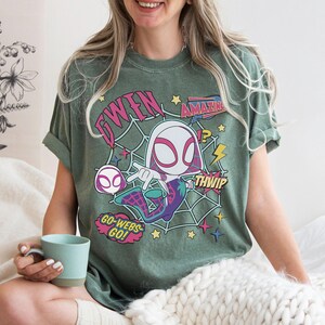 May include: A sage green short-sleeve t-shirt featuring a graphic of a cartoon character in a white and pink hooded suit. The design includes a spider web background, yellow stars, and text that reads GWEN, AMAZING, THWIP, and GO-WEBS-GO.