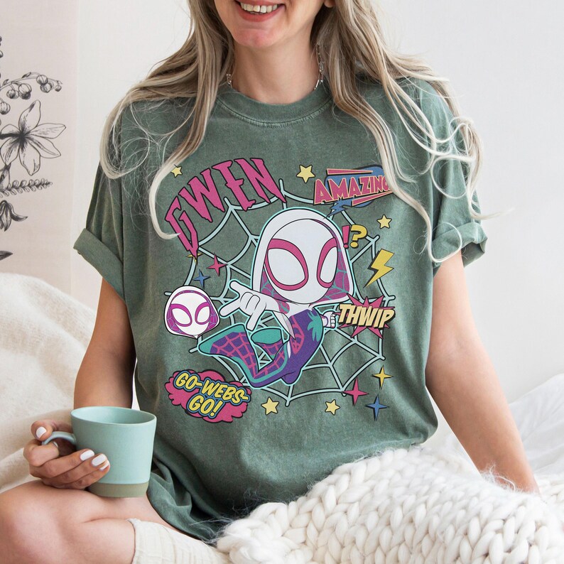 May include: A sage green short-sleeve t-shirt featuring a graphic of a cartoon character in a white and pink hooded suit. The design includes a spider web background, yellow stars, and text that reads GWEN, AMAZING, THWIP, and GO-WEBS-GO.