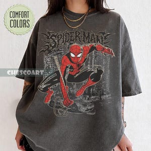 May include: Dark gray t-shirt featuring a red and black Spider-Man graphic. The design includes the word "SPIDERMAN" in a stylized font. The shirt also has the text "COMFORT COLORS" and "CHESCOART" on it.
