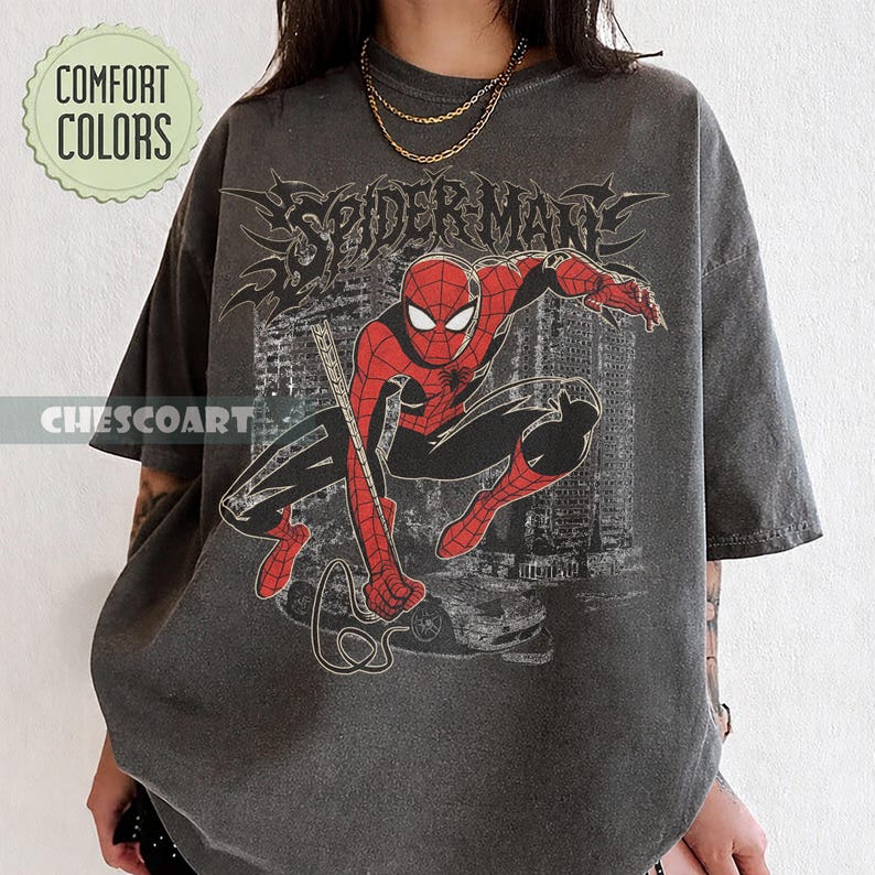 May include: Dark gray t-shirt featuring a red and black Spider-Man graphic. The design includes the word "SPIDERMAN" in a stylized font. The shirt also has the text "COMFORT COLORS" and "CHESCOART" on it.