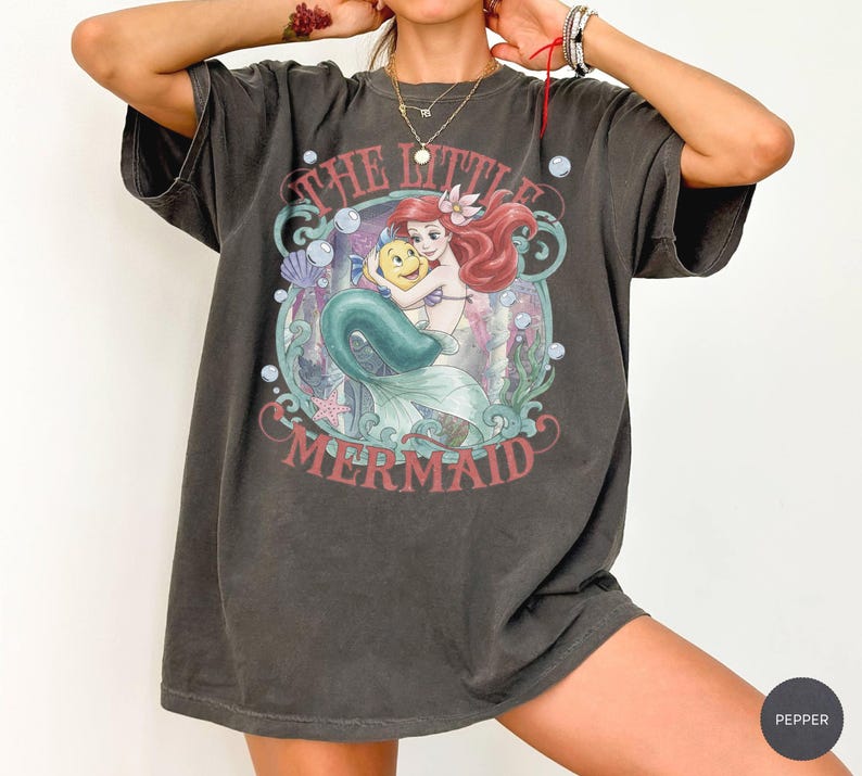 May include: A charcoal gray short-sleeve t-shirt featuring a graphic print of Ariel and Flounder from The Little Mermaid. The design includes underwater elements like bubbles, seaweed, and seashells, with the text The Little Mermaid in red.