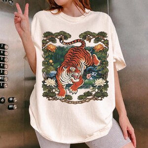 K&ouml;nnte beinhalten: Cremefarbenes T-Shirt mit einer detaillierten Illustration eines orange-schwarzen Tigers, umrahmt von Blumen und Bl&auml;ttern. Der Tiger ist das zentrale Motiv, mit einem Landschaftshintergrund.