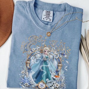 May include: A blue Comfort Colors t-shirt featuring a graphic print of Elsa and Olaf from Frozen. The design includes the name Elsa in gold lettering, a decorative archway, and snowflake accents. A gold-toned necklace with a small rose pendant rests on the collar.
