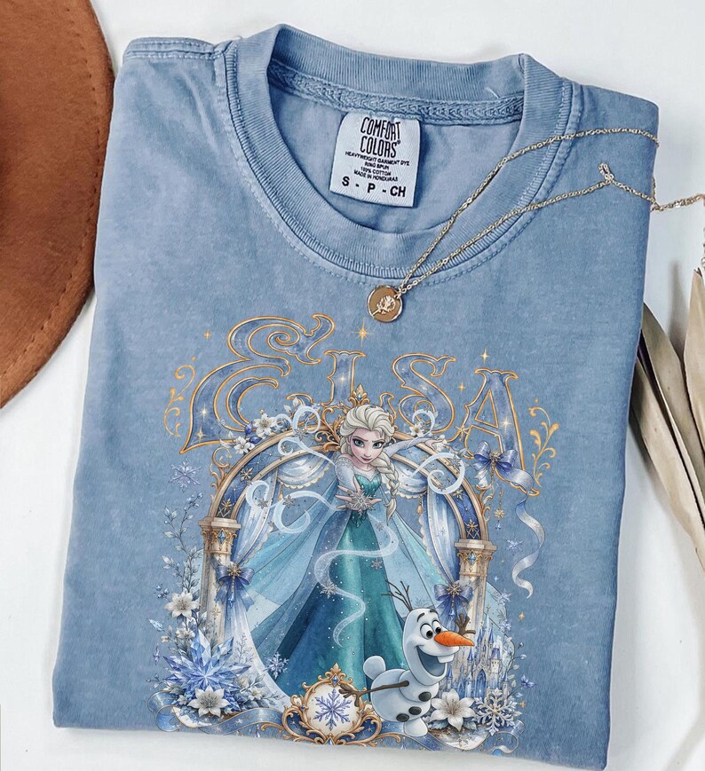 May include: A blue Comfort Colors t-shirt featuring a graphic print of Elsa and Olaf from Frozen. The design includes the name Elsa in gold lettering, a decorative archway, and snowflake accents. A gold-toned necklace with a small rose pendant rests on the collar.