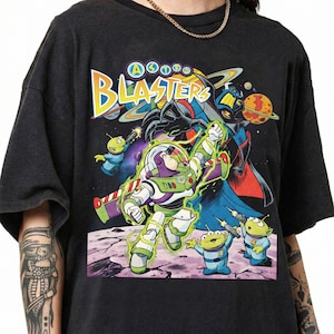 Puede incluir: Una camiseta negra de manga corta con un estampado gr&aacute;fico colorido de Buzz Lightyear y alien&iacute;genas verdes de Toy Story. El texto "ASTRO BLASTERS" est&aacute; impreso en amarillo y blanco en la parte superior, con un fondo espacial y planetas.