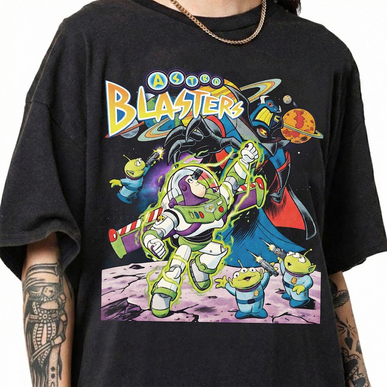 Puede incluir: Una camiseta negra de manga corta con un estampado gr&aacute;fico colorido de Buzz Lightyear y alien&iacute;genas verdes de Toy Story. El texto "ASTRO BLASTERS" est&aacute; impreso en amarillo y blanco en la parte superior, con un fondo espacial y planetas.