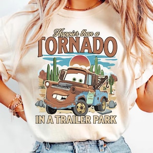 May include: Cream-coloured t-shirt featuring a cartoon tow truck in a desert landscape with cacti and a setting sun. The text reads "Happier than a TORNADO IN A TRAILER PARK" in brown and black lettering.