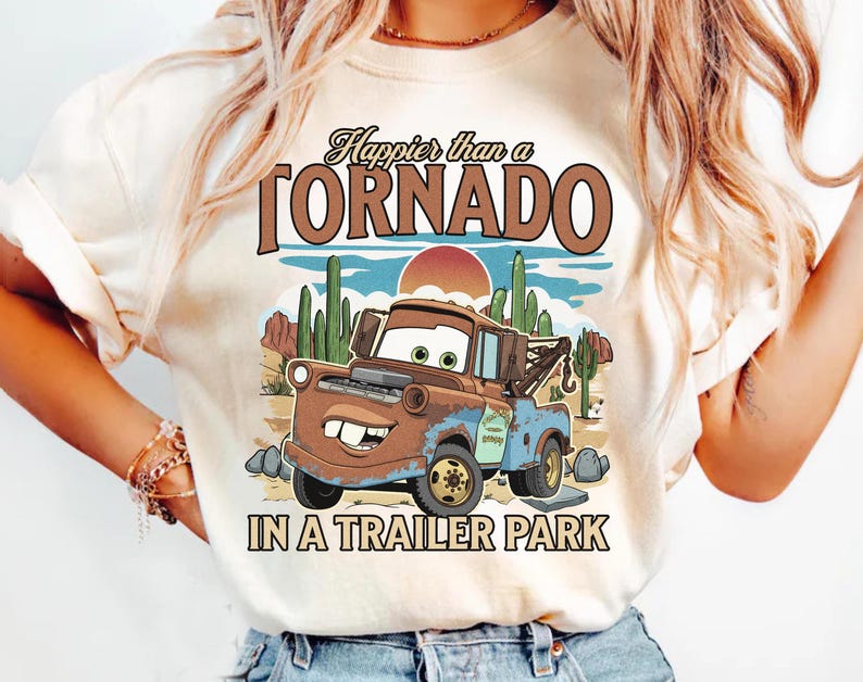 May include: Cream-coloured t-shirt featuring a cartoon tow truck in a desert landscape with cacti and a setting sun. The text reads "Happier than a TORNADO IN A TRAILER PARK" in brown and black lettering.