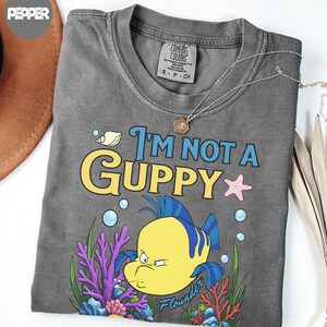 Puede incluir: Camiseta gris con el texto "I'm Not a Guppy" y una ilustraci&oacute;n de dibujos animados de un pez amarillo llamado Flounder. El dise&ntilde;o incluye burbujas azules, una estrella de mar rosa y coral.