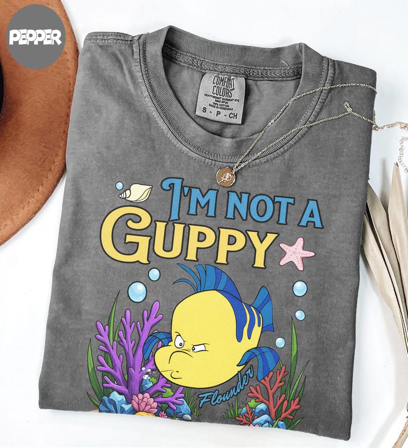 Puede incluir: Camiseta gris con el texto "I'm Not a Guppy" y una ilustraci&oacute;n de dibujos animados de un pez amarillo llamado Flounder. El dise&ntilde;o incluye burbujas azules, una estrella de mar rosa y coral.